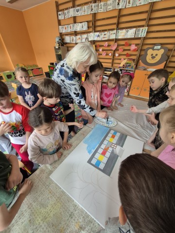 Na obrazku widać grupę dzieci malującą wraz z Panią prowadzącą warsztaty malarskie.